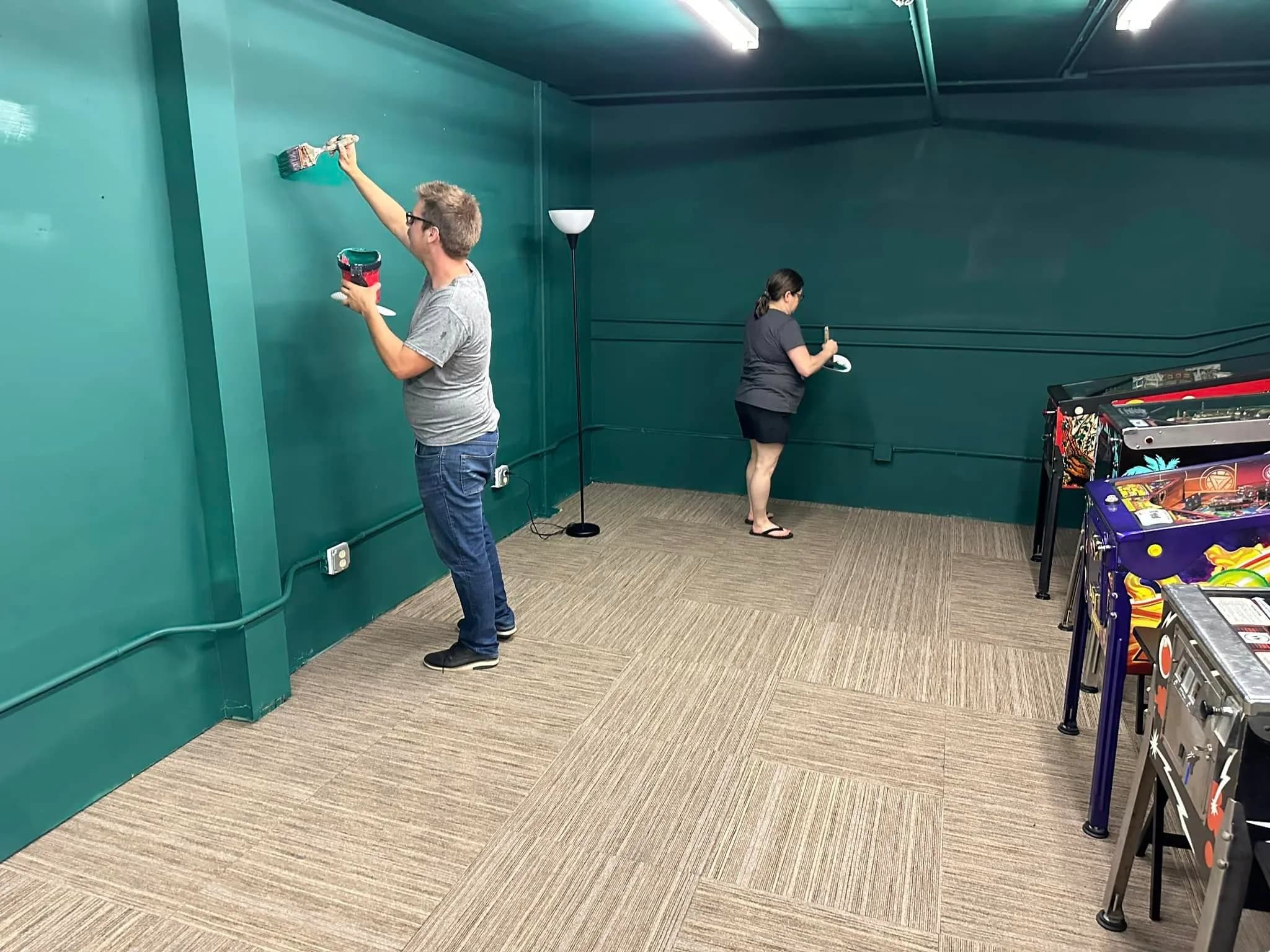 Two people painting teal walls in a room with pinball machines