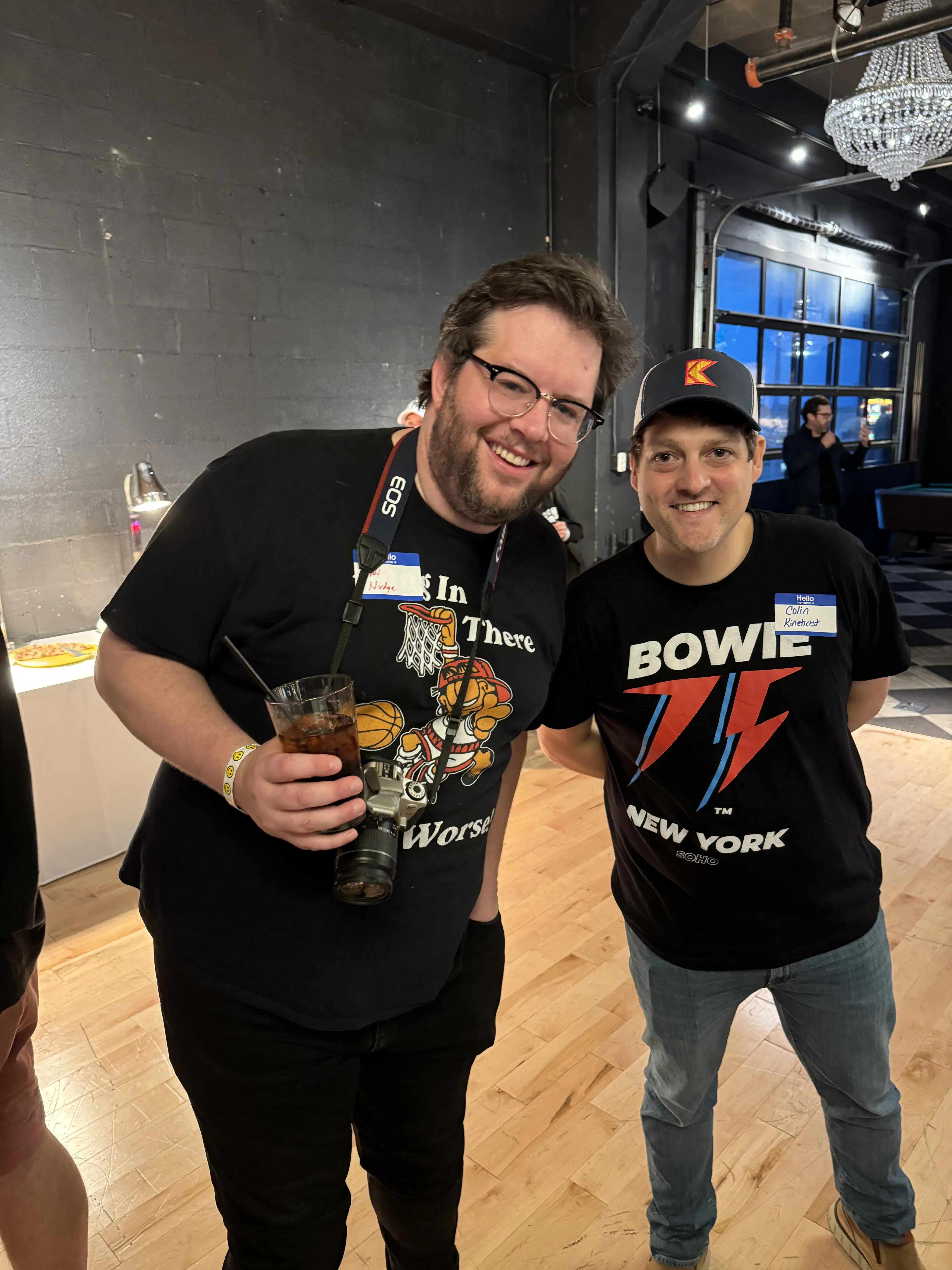 Two men in graphic tees smiling at event with drinks and name tags