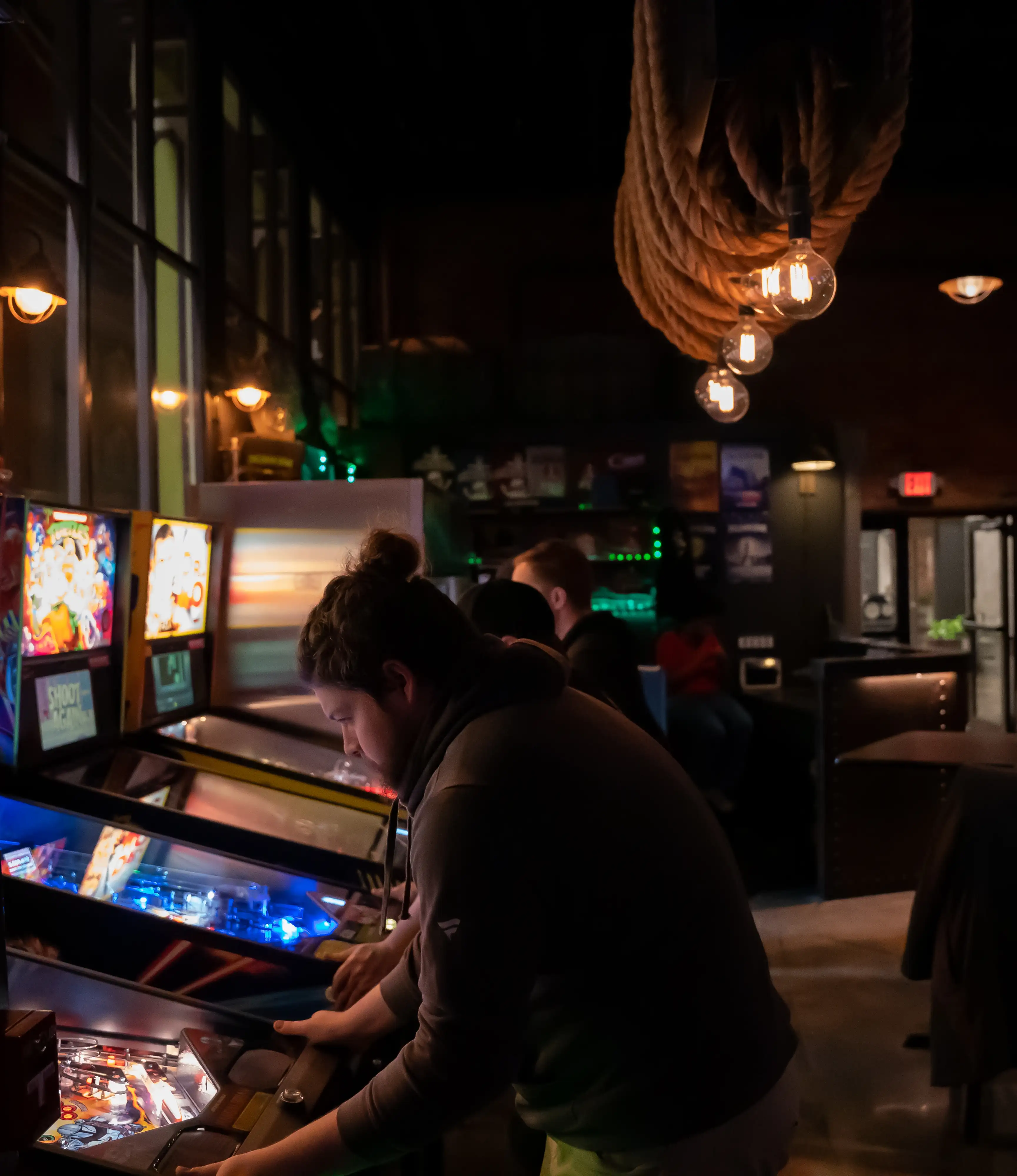 Dimly lit arcade with people playing pinball machines under decorative lights