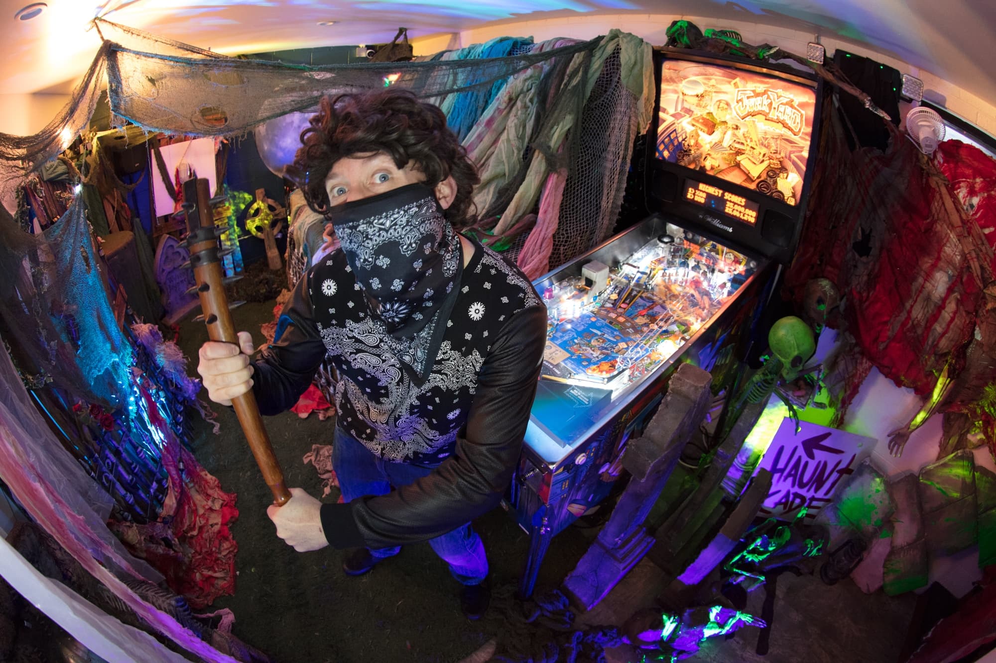 Masked person with staff near colorful pinball machine in eccentric, draped space