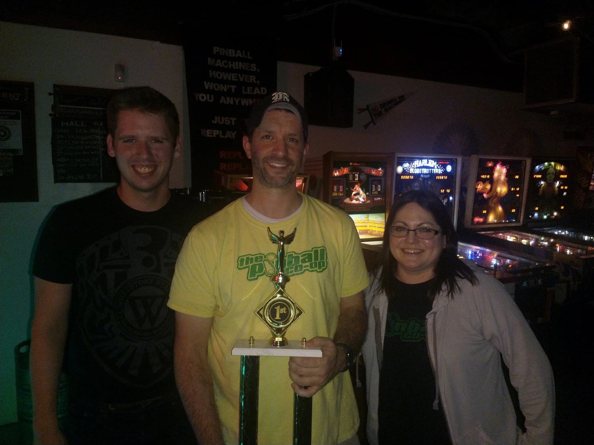 Three people posing with a first-place trophy at a pinball arcade