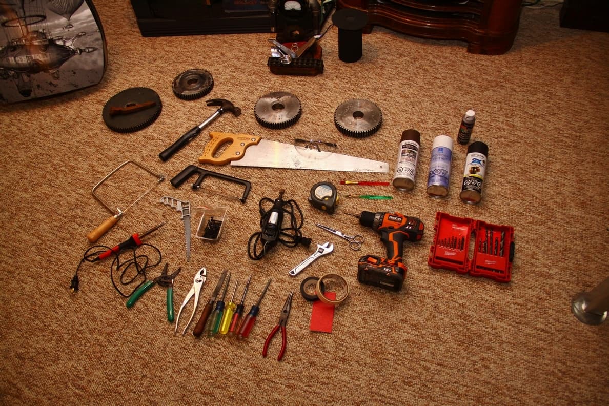 Assorted workshop tools spread out on carpeted floor, including saw, drill, gears