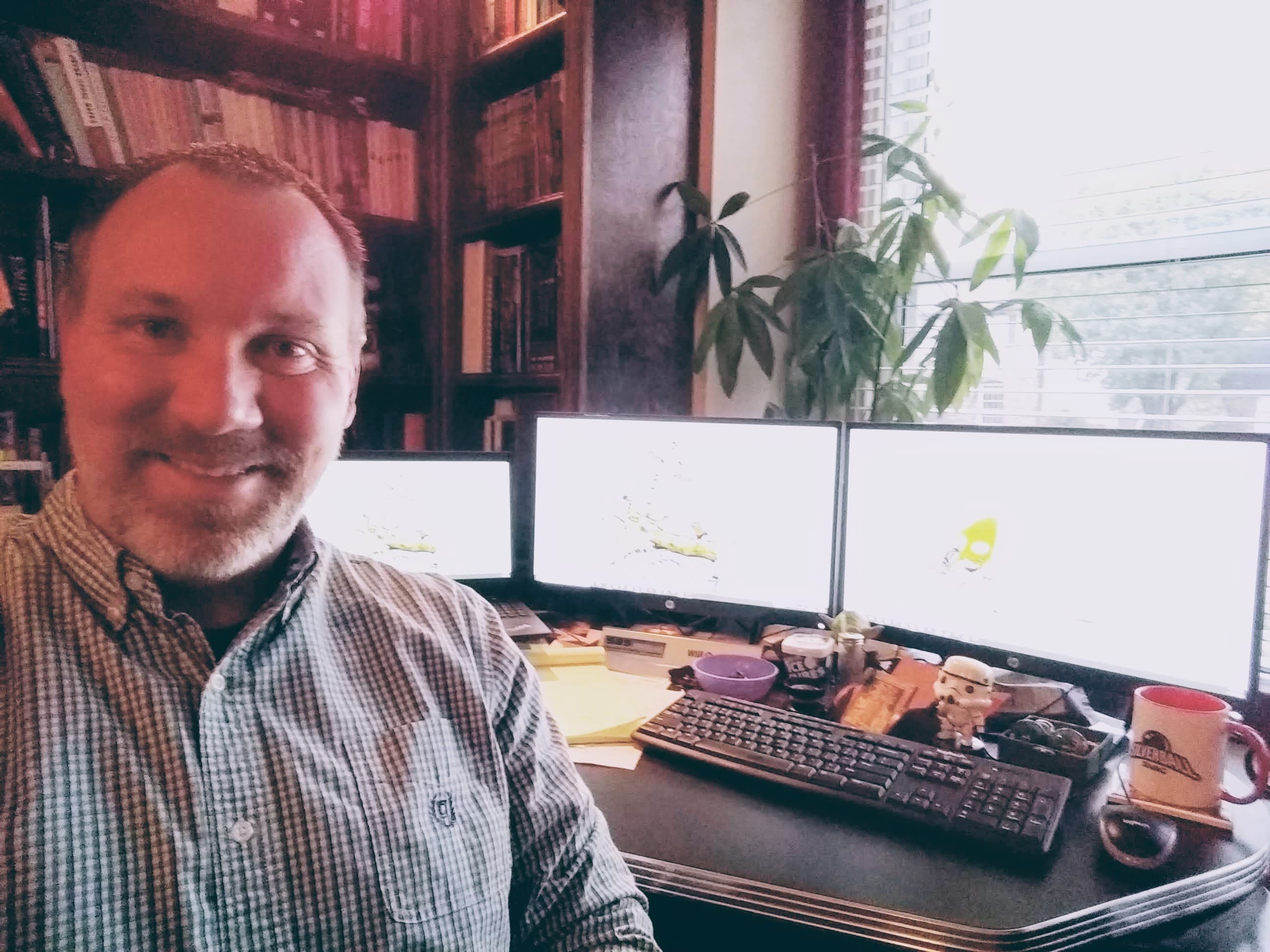 Smiling person at workspace with multiple monitors, bookshelves, and plants