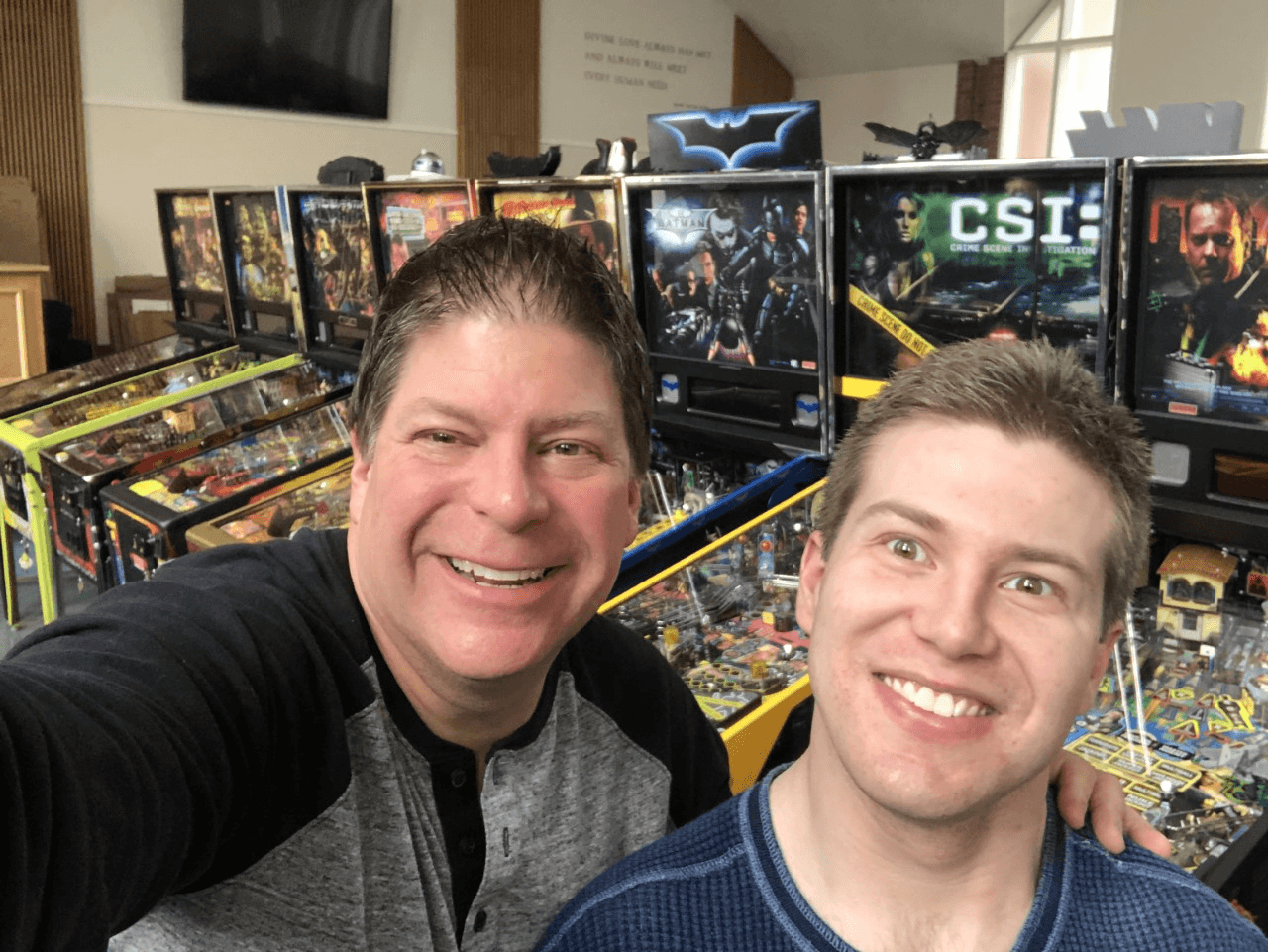 Two men smiling in front of pinball machines and pop culture posters